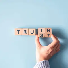 Wooden blocks spell "TRUST". A hand is placing the final block, symbolizing how to be more authentic by building trust.