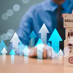 Building skills for job security. A hand stacks wooden blocks with icons representing learning, teamwork, and goals, amidst rising arrows.