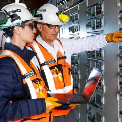 Two engineers in a control room, one pointing at a panel, the other holding a laptop, showcasing the characteristics of innovation in electrical systems.