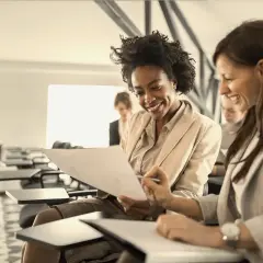 Two women smile while reviewing documents in a classroom setting, likely participating in leadership courses for women.