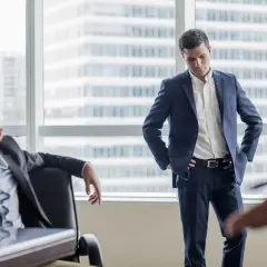 A man in a suit looks down, head in hand, as another man stands with hands on hips. This scene illustrates how to deliver bad news with empathy.