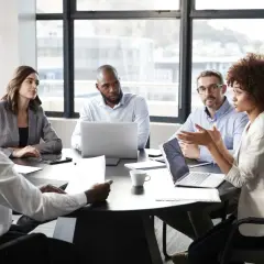 A diverse group of professionals engaged in a lively meeting discussion around a table, with laptops and papers spread out.