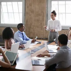 A diverse group attends a board of directors training program. A woman in a white shirt leads the session, while others engage with laptops and tablets.
