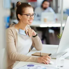 A smiling woman in glasses works at a computer, holding a phone.