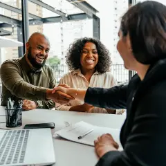 A man and woman smile, shaking hands after successful negotiation classes online. A laptop shows charts.