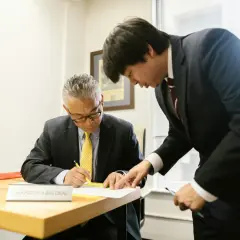 Two men in suits review documents. One man, seated, signs papers while the other, standing, points to the document. This scene prompts questions to ask your boss.