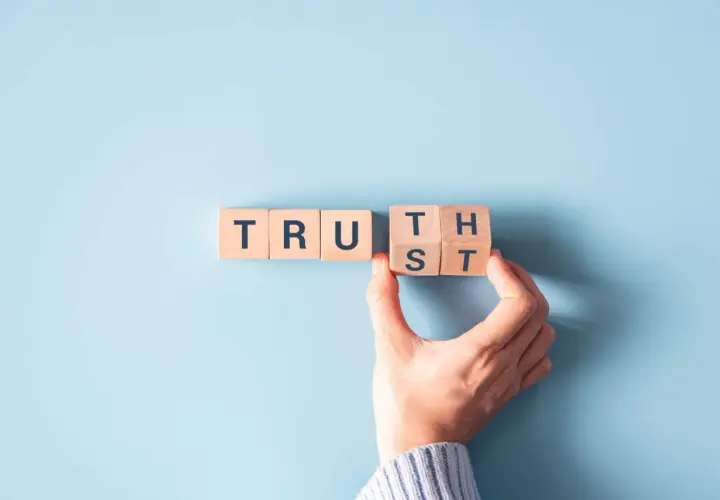 Wooden blocks spell "TRUST". A hand is placing the final block, symbolizing how to be more authentic by building trust.