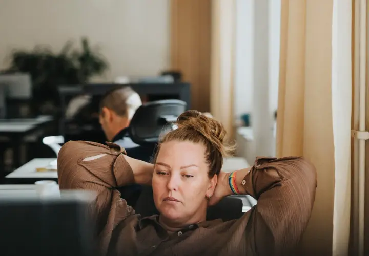 A woman with her hands behind her head, looking tired at her desk. This image captures the essence of presenteeism, where someone is physically at work but not fully engaged or productive.