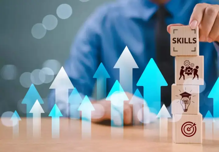 Building skills for job security. A hand stacks wooden blocks with icons representing learning, teamwork, and goals, amidst rising arrows.