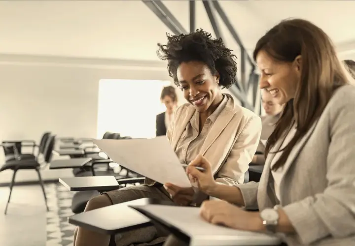 Two women smile while reviewing documents in a classroom setting, likely participating in leadership courses for women.