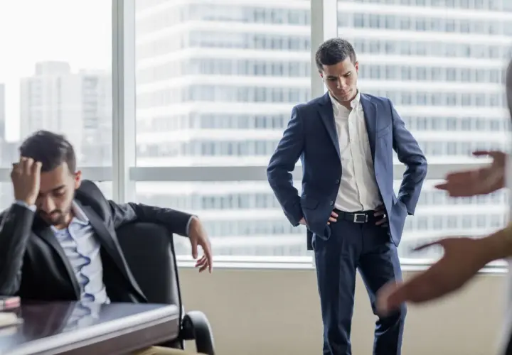 A man in a suit looks down, head in hand, as another man stands with hands on hips. This scene illustrates how to deliver bad news with empathy.