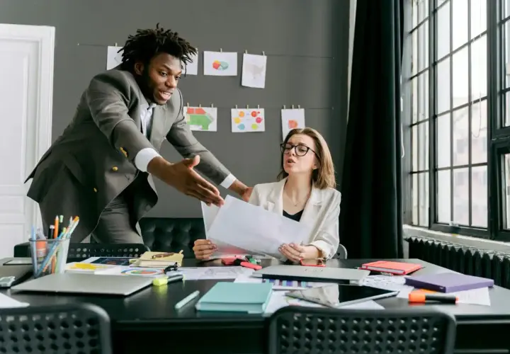 A man in a suit gestures emphatically during a difficult conversations training session with a woman at a desk.