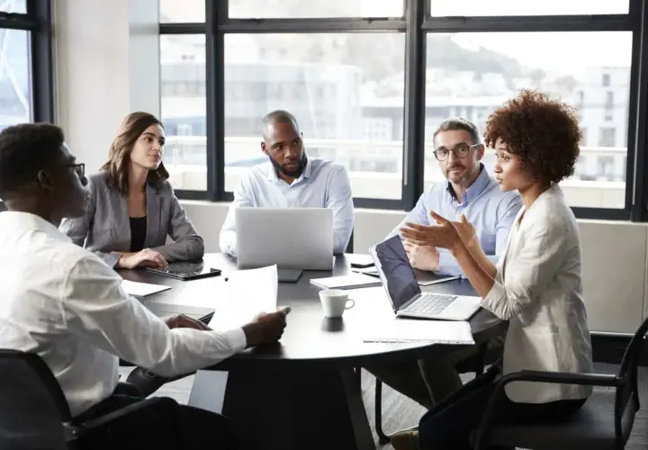 A diverse group of professionals engaged in a lively meeting discussion around a table, with laptops and papers spread out.