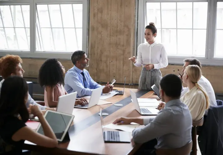 A diverse group attends a board of directors training program. A woman in a white shirt leads the session, while others engage with laptops and tablets.
