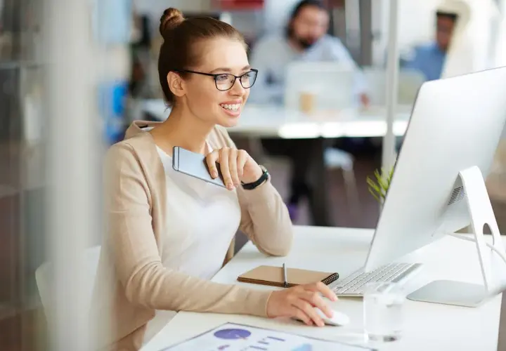 A smiling woman in glasses works at a computer, holding a phone.