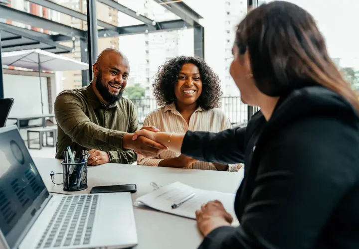A man and woman smile, shaking hands after successful negotiation classes online. A laptop shows charts.