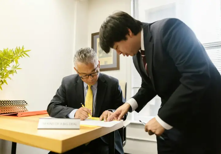Two men in suits review documents. One man, seated, signs papers while the other, standing, points to the document. This scene prompts questions to ask your boss.