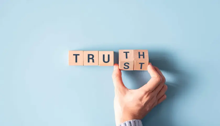 Wooden blocks spell "TRUST". A hand is placing the final block, symbolizing how to be more authentic by building trust.