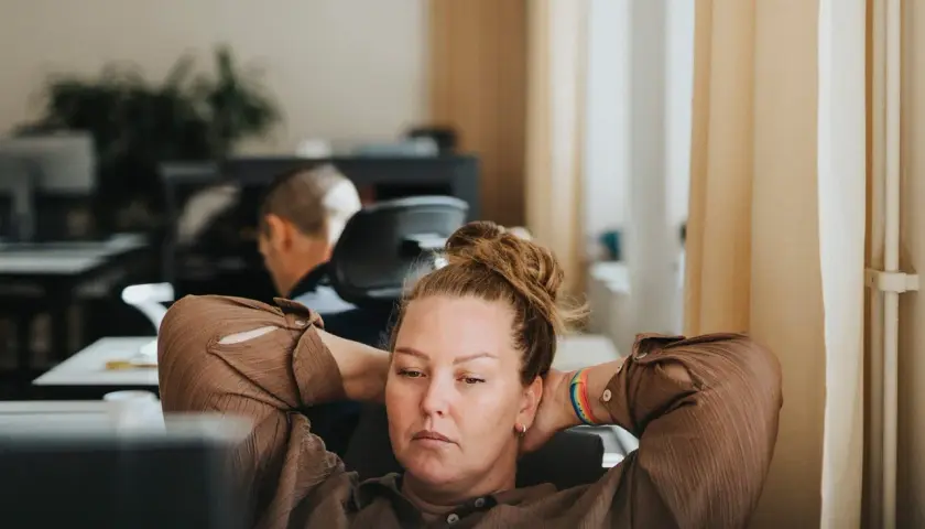 A woman with her hands behind her head, looking tired at her desk. This image captures the essence of presenteeism, where someone is physically at work but not fully engaged or productive.