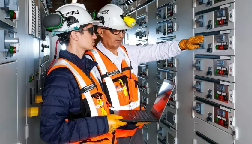 Two engineers in a control room, one pointing at a panel, the other holding a laptop, showcasing the characteristics of innovation in electrical systems.