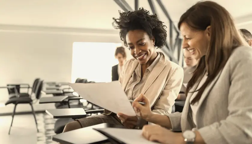 Two women smile while reviewing documents in a classroom setting, likely participating in leadership courses for women.