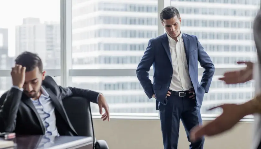 A man in a suit looks down, head in hand, as another man stands with hands on hips. This scene illustrates how to deliver bad news with empathy.