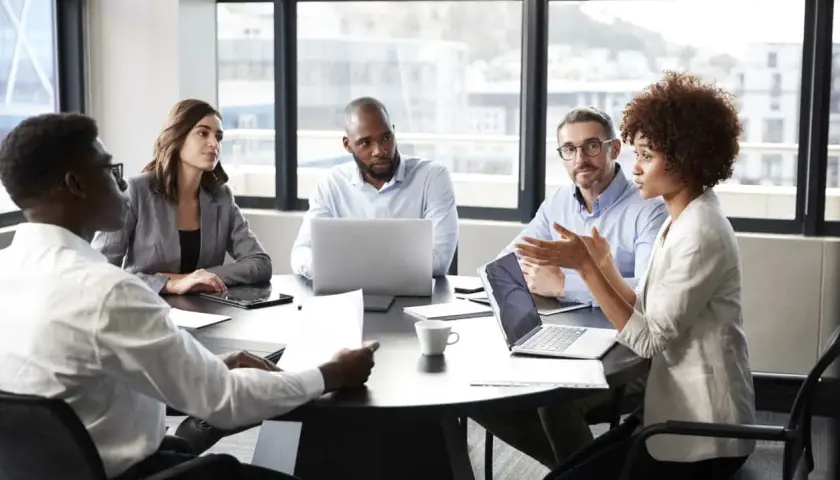 A diverse group of professionals engaged in a lively meeting discussion around a table, with laptops and papers spread out.