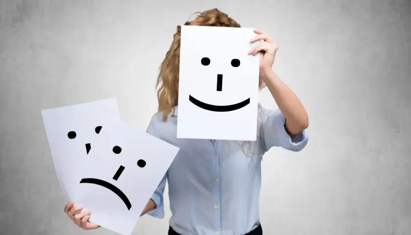 A person holds a happy face, while another sad face is shown. This could represent people who complain all the time, hiding their true feelings.