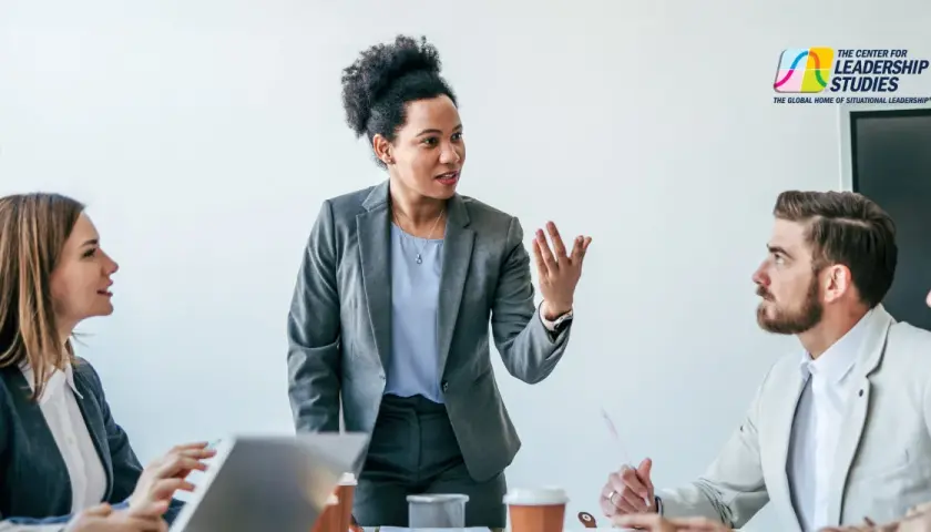 A diverse team engages in a communications leadership development program, with a woman in a grey suit leading the discussion.