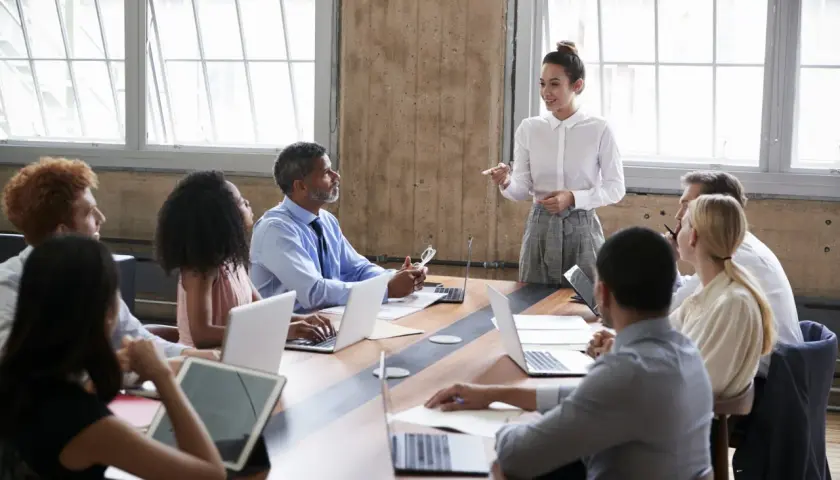 A diverse group attends a board of directors training program. A woman in a white shirt leads the session, while others engage with laptops and tablets.