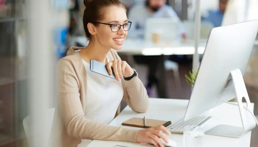 A smiling woman in glasses works at a computer, holding a phone.