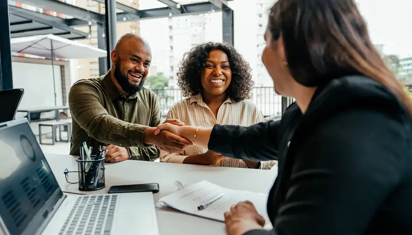 A man and woman smile, shaking hands after successful negotiation classes online. A laptop shows charts.