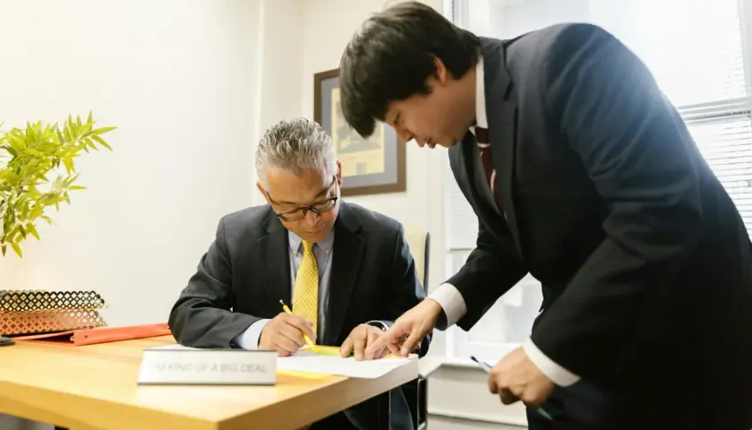 Two men in suits review documents. One man, seated, signs papers while the other, standing, points to the document. This scene prompts questions to ask your boss.