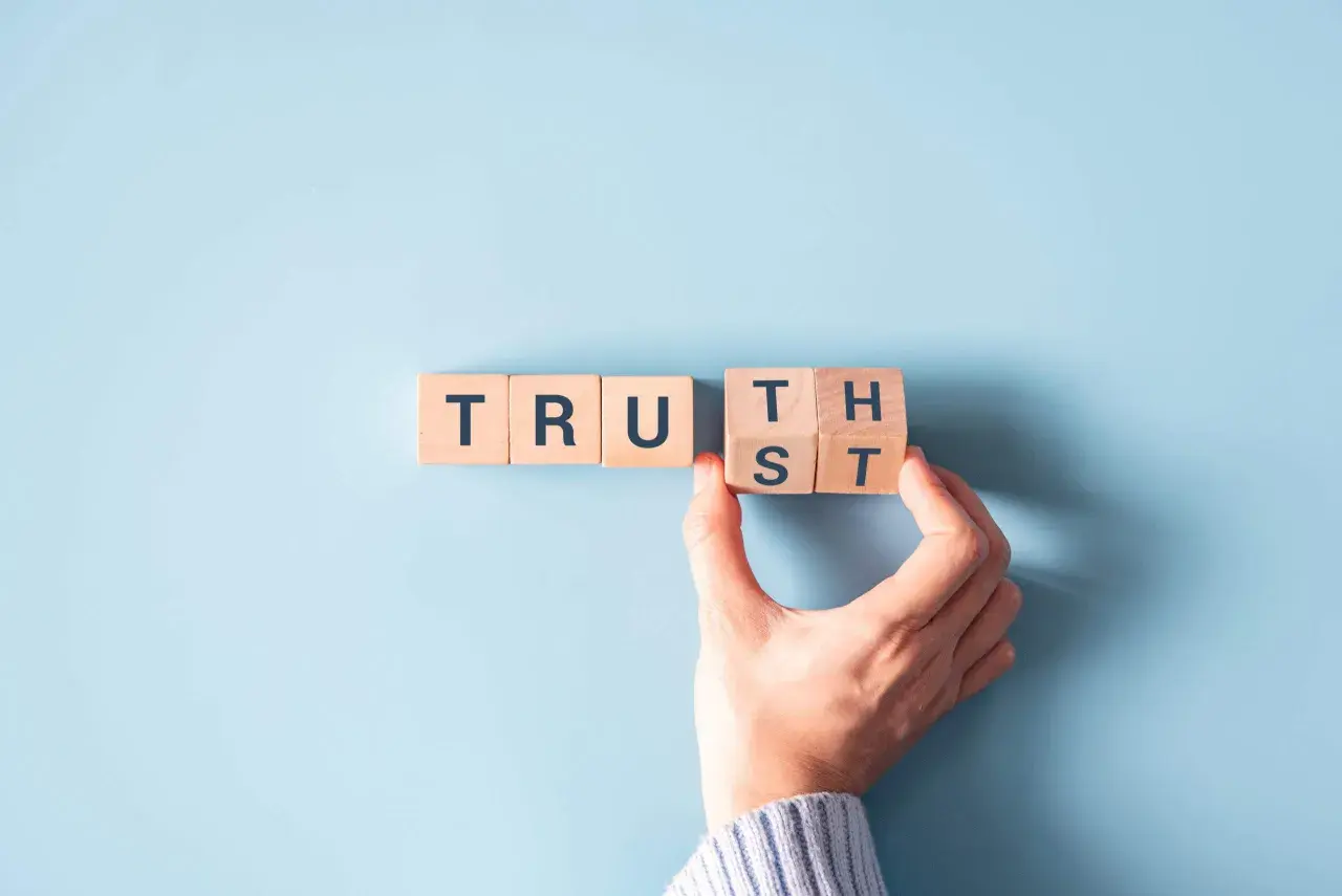 Wooden blocks spell "TRUST". A hand is placing the final block, symbolizing how to be more authentic by building trust.