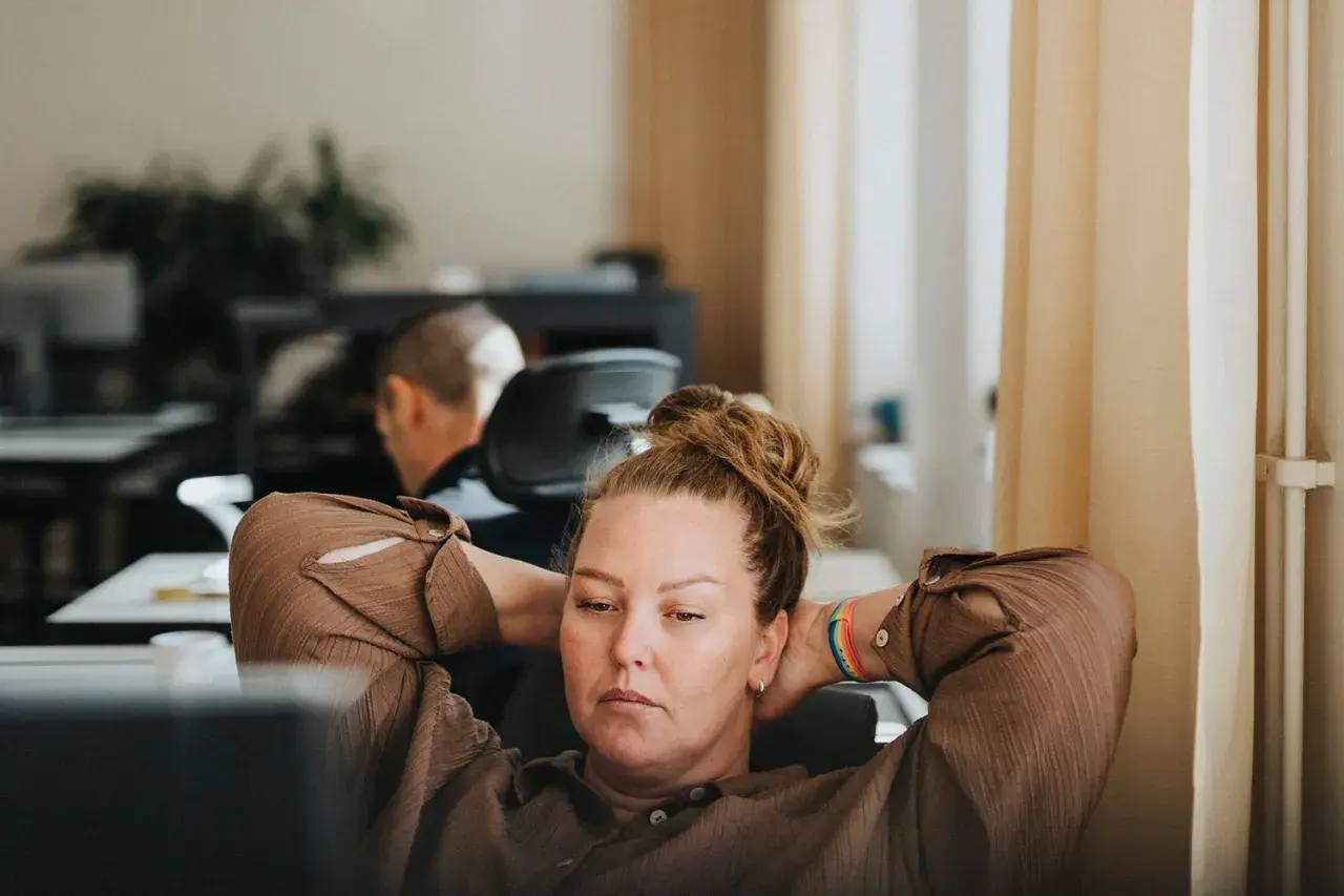 A woman with her hands behind her head, looking tired at her desk. This image captures the essence of presenteeism, where someone is physically at work but not fully engaged or productive.