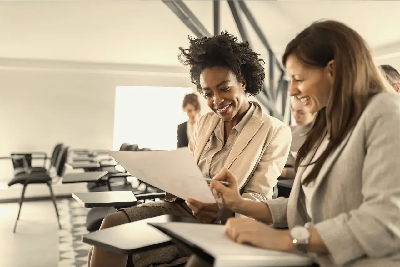 Two women smile while reviewing documents in a classroom setting, likely participating in leadership courses for women.
