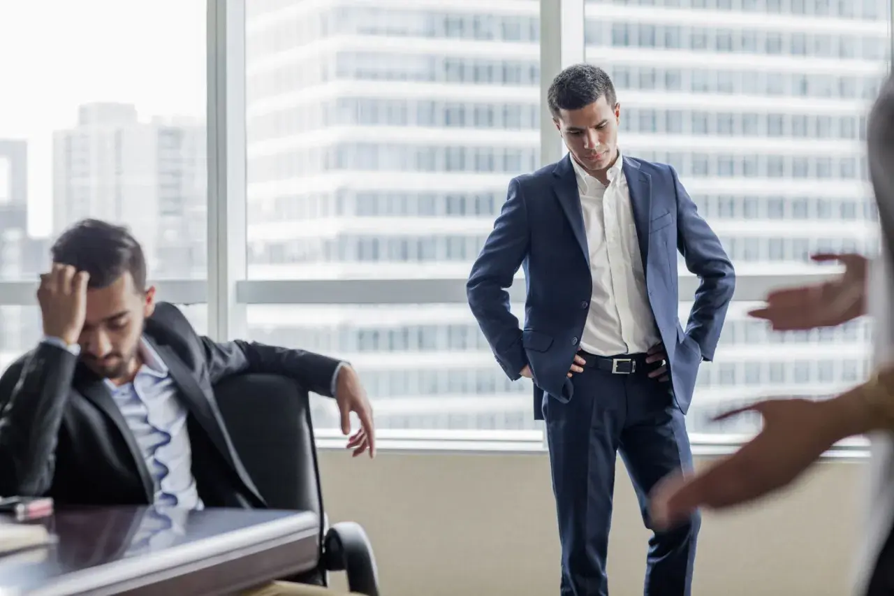 A man in a suit looks down, head in hand, as another man stands with hands on hips. This scene illustrates how to deliver bad news with empathy.