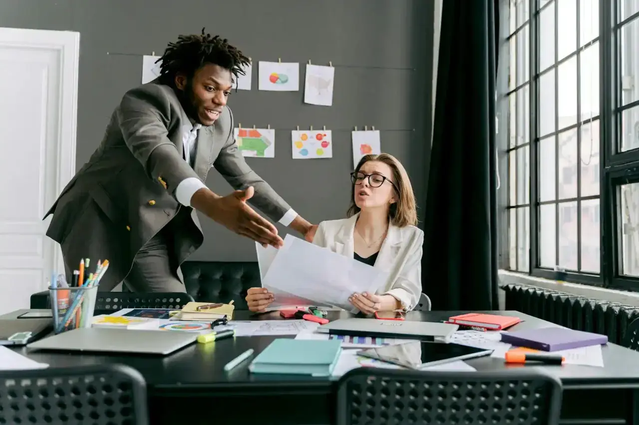 A man in a suit gestures emphatically during a difficult conversations training session with a woman at a desk.