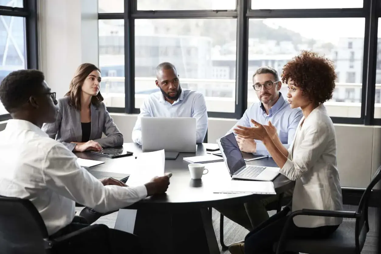 A diverse group of professionals engaged in a lively meeting discussion around a table, with laptops and papers spread out.