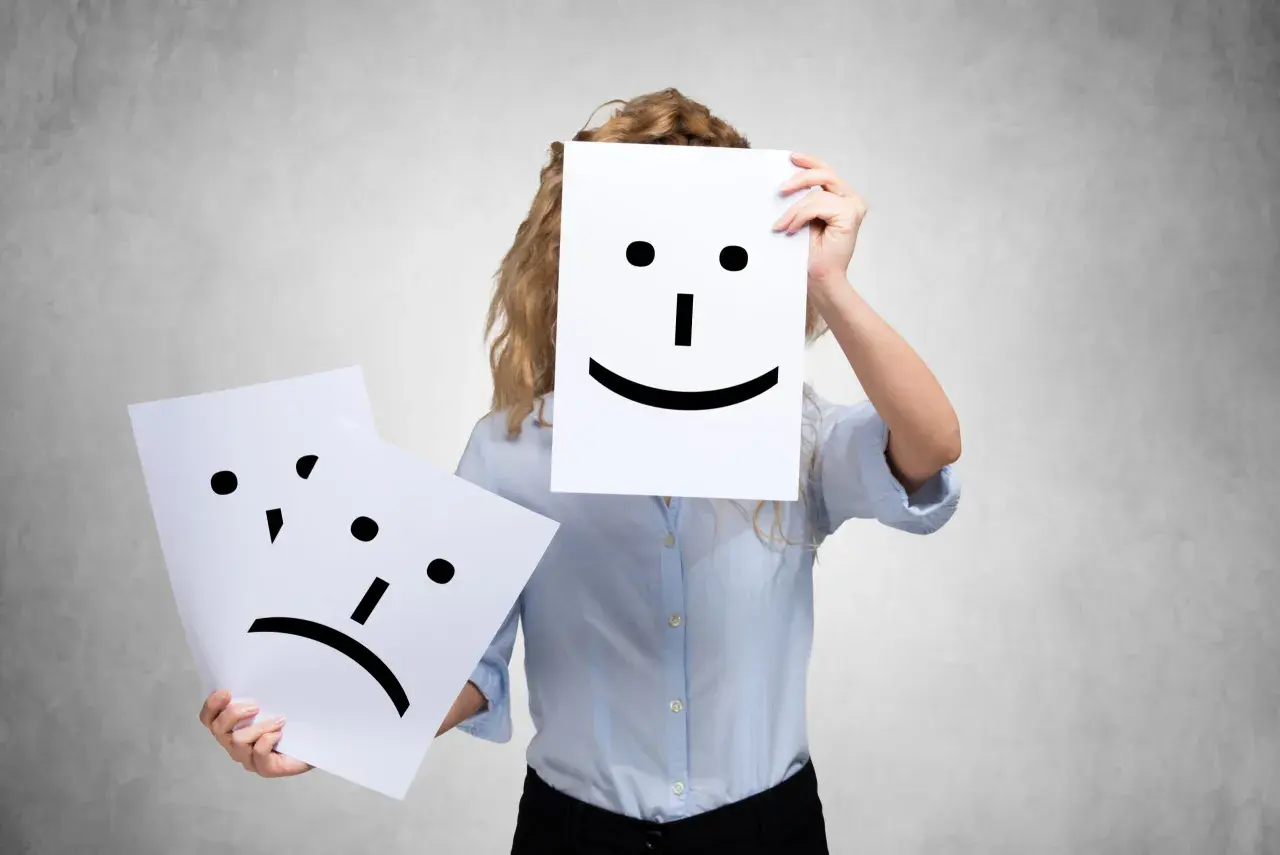 A person holds a happy face, while another sad face is shown. This could represent people who complain all the time, hiding their true feelings.