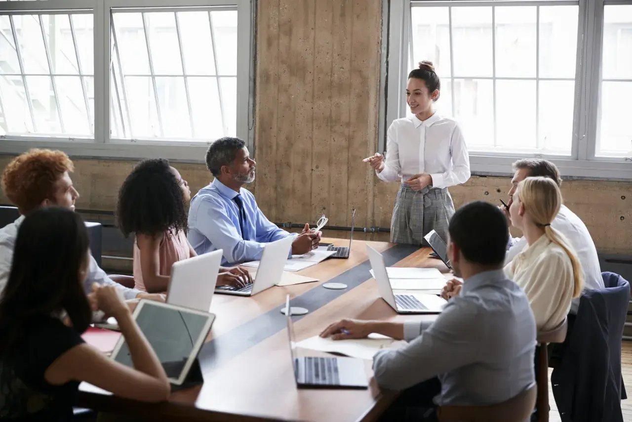 A diverse group attends a board of directors training program. A woman in a white shirt leads the session, while others engage with laptops and tablets.