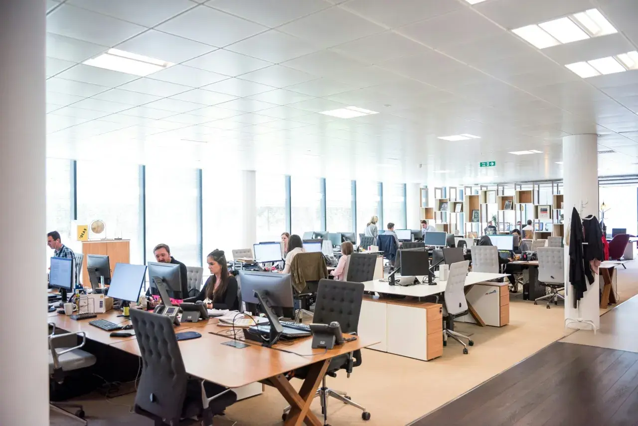 A busy open office space with employees working at desks. Natural light streams in from large windows, illuminating the modern workstations.
