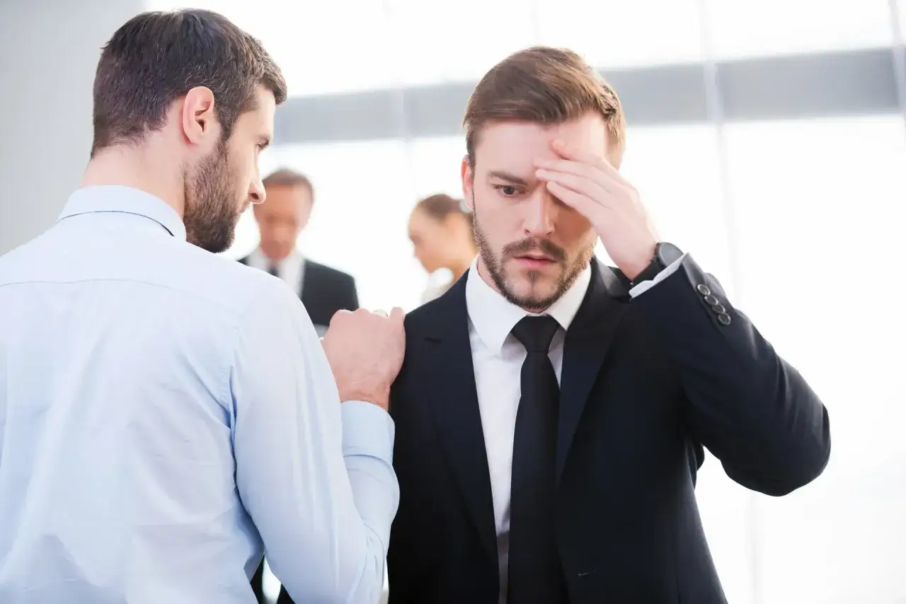 A man in a suit looks distressed, hand on his forehead. Another man in a light blue shirt places a comforting hand on his shoulder, demonstrating how to deliver bad news with empathy.