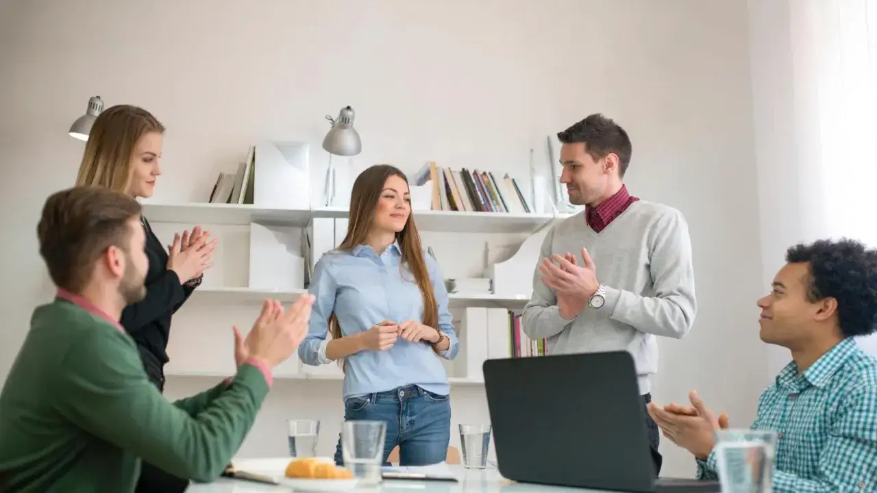 A team applauds a colleague who is about to introduce myself. They are in a bright office with bookshelves in the background.