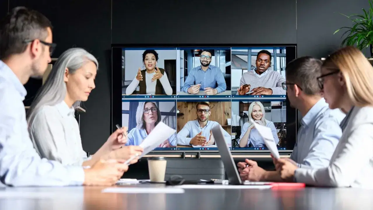 A diverse group of professionals engage in a lively meeting discussion via video conference, with some participants in the foreground and others on the large screen.