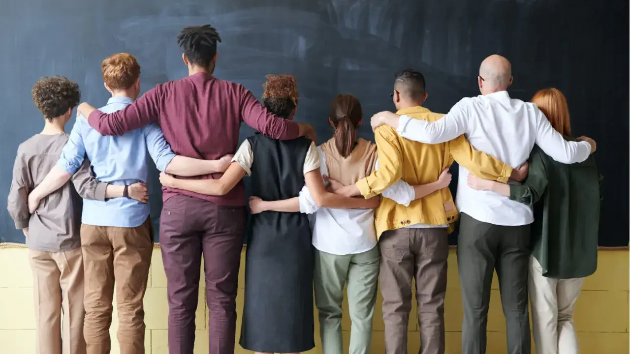 A diverse group stands with arms around each other, a symbol of strong leadership and mentoring, united against a blackboard.