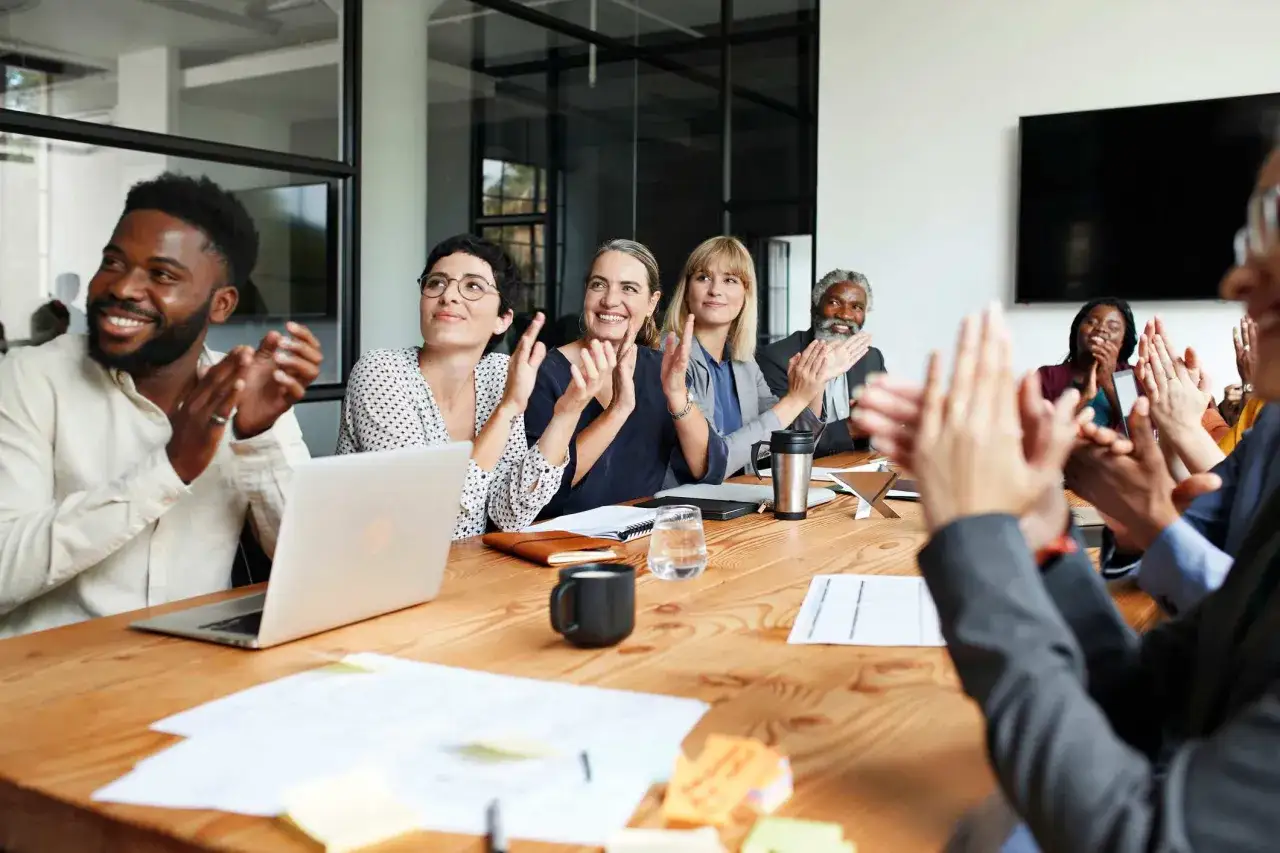 A diverse group of colleagues are clapping, celebrating a success. One man smiles, asking for help with a project.
