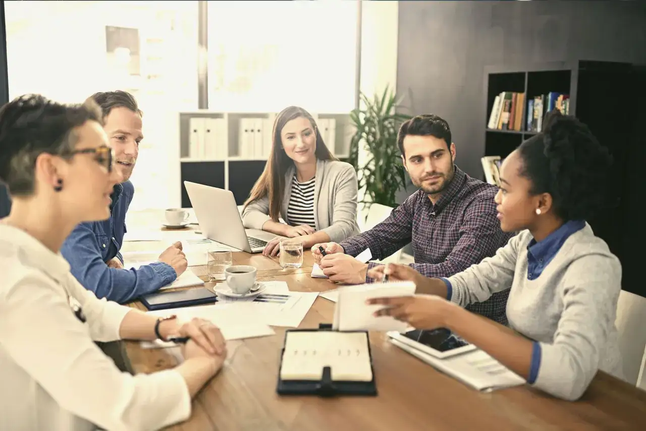 A diverse team collaborates around a table, with a positive tone at the top. They're engaged in discussion, with laptops and papers spread out.