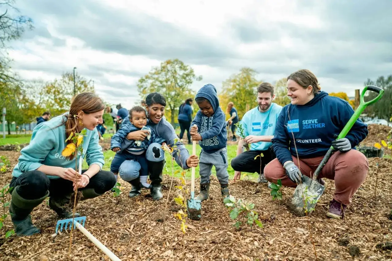 A diverse group, including children, participates in corporate volunteering, planting saplings. Everyone is smiling and engaged in the community gardening effort.