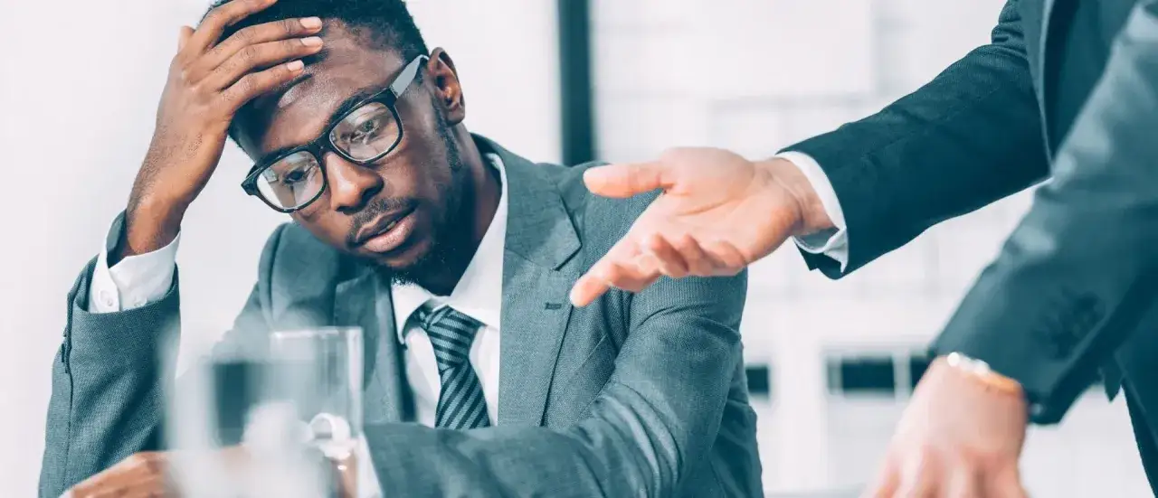 A stressed Black man in a suit with glasses holds his head, wondering, 